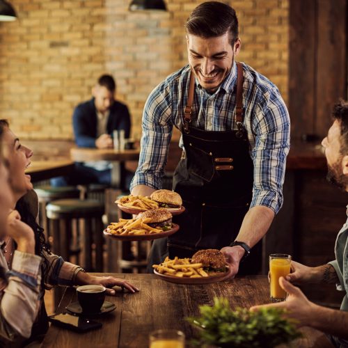 Group of happy friends having fun while waiter is serving them food in a pub. Focus is on waiter.